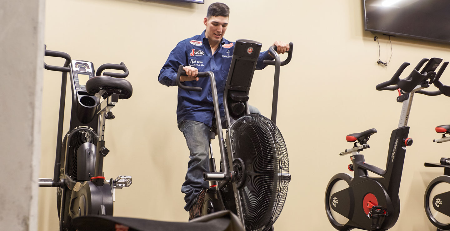 Jess Pope is wearing a blue button-down shirt and blue jeans, riding a stationary bike to warm up before competing at a rodeo. 